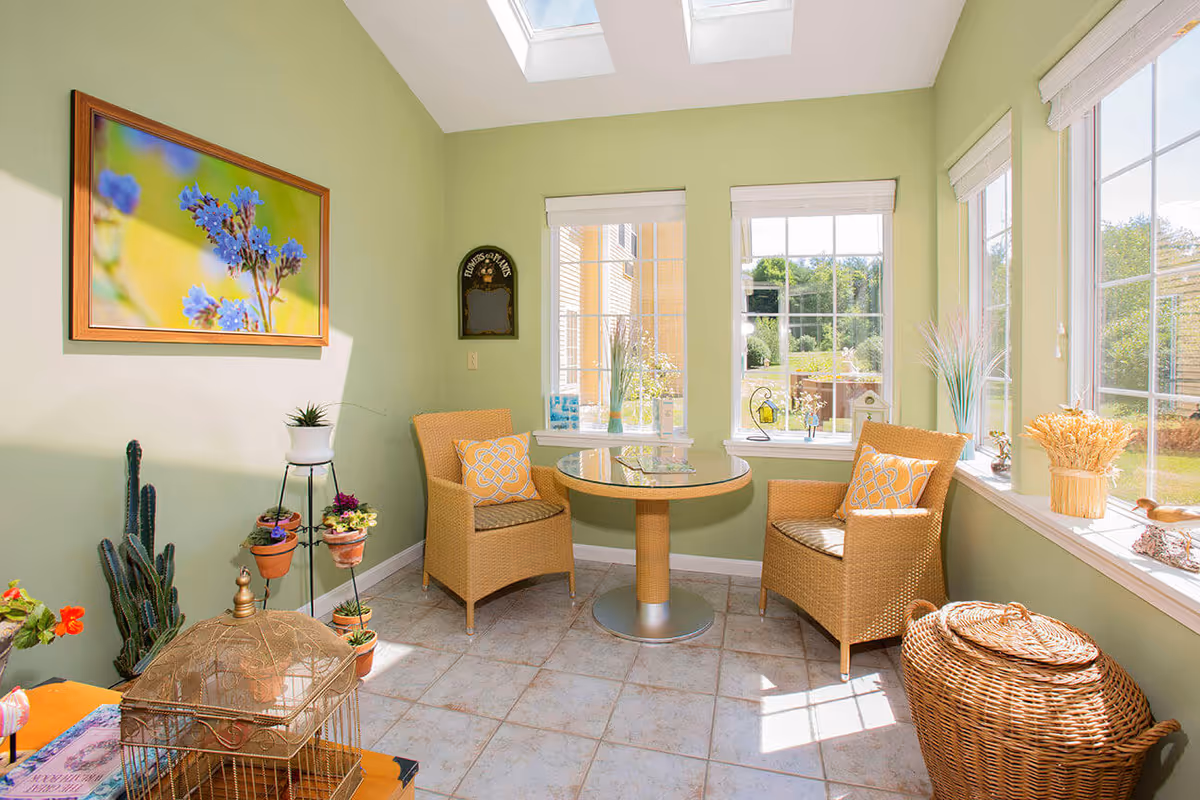 Sunlit green sunroom with skylights, two wicker chairs around a small round table and plants by large windows.