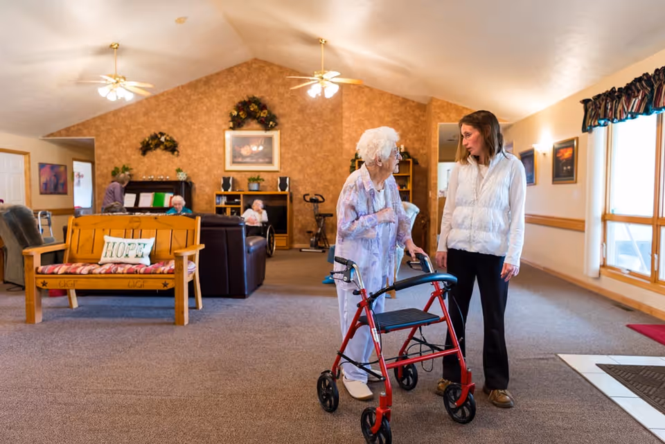 An elderly woman using a red walker is talking to a younger woman in a white vest inside a spacious living room area of an assisted living facility. The room has beige walls, carpeted floors, ceiling fans, and large windows with curtains. In the background, other elderly residents are seated on sofas and chairs near a fireplace and a piano.