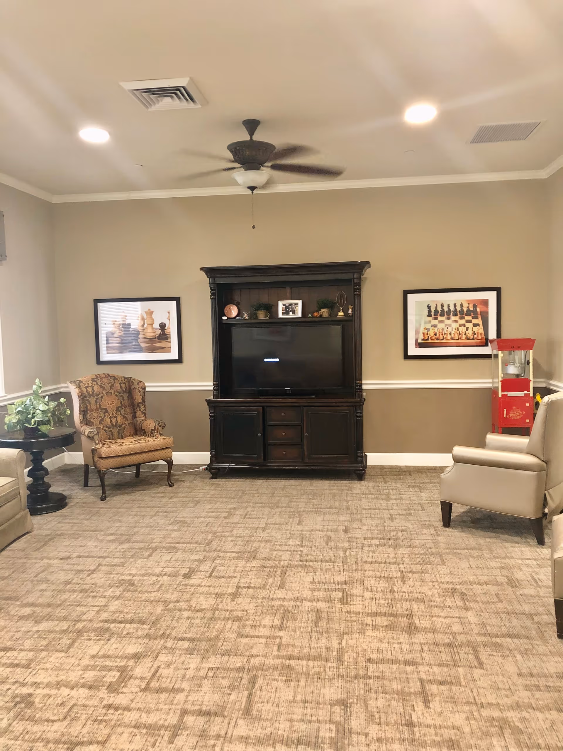 A cozy living room with a ceiling fan, a dark wooden entertainment center holding a TV, two framed chess-themed pictures on the wall, a patterned armchair, a beige armchair, a small round table with a plant, and a red popcorn machine in the corner.