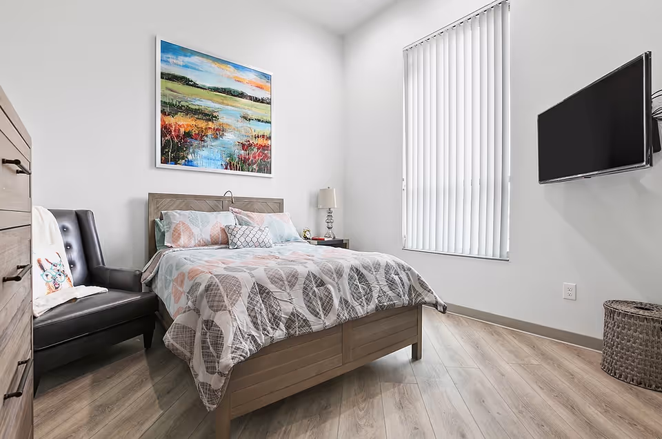 A bright bedroom with a wooden bed featuring a patterned comforter and pillows, a black leather chair with a decorative pillow, a wooden dresser, a nightstand with a lamp, a large window with vertical blinds, a wall-mounted flat screen TV, and a colorful landscape painting above the bed.
