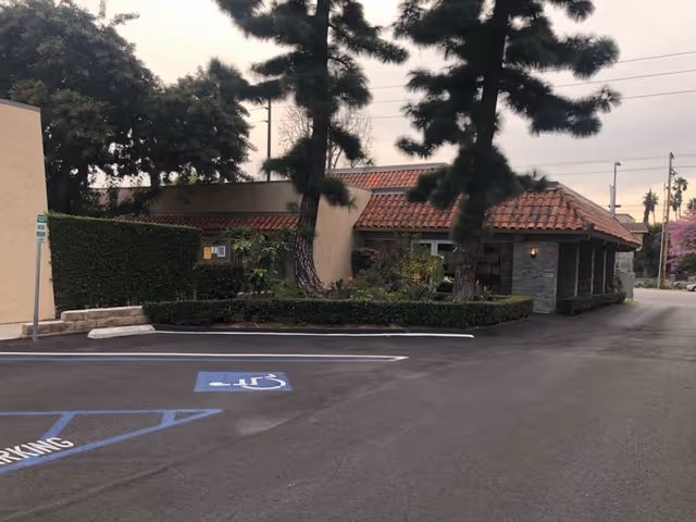 Parking lot and entrance of a single-story building with a red tile roof, tall pine trees, and a marked handicapped parking space.
