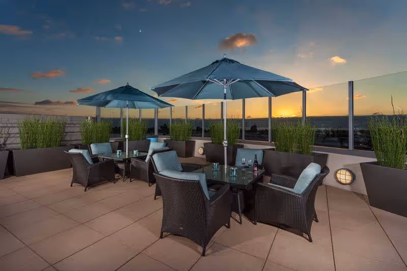 Outdoor patio area at sunset with two tables, each surrounded by four wicker chairs with cushions and shaded by large blue umbrellas. Tall green plants in rectangular planters line the perimeter, and a glass railing offers a view of the horizon.
