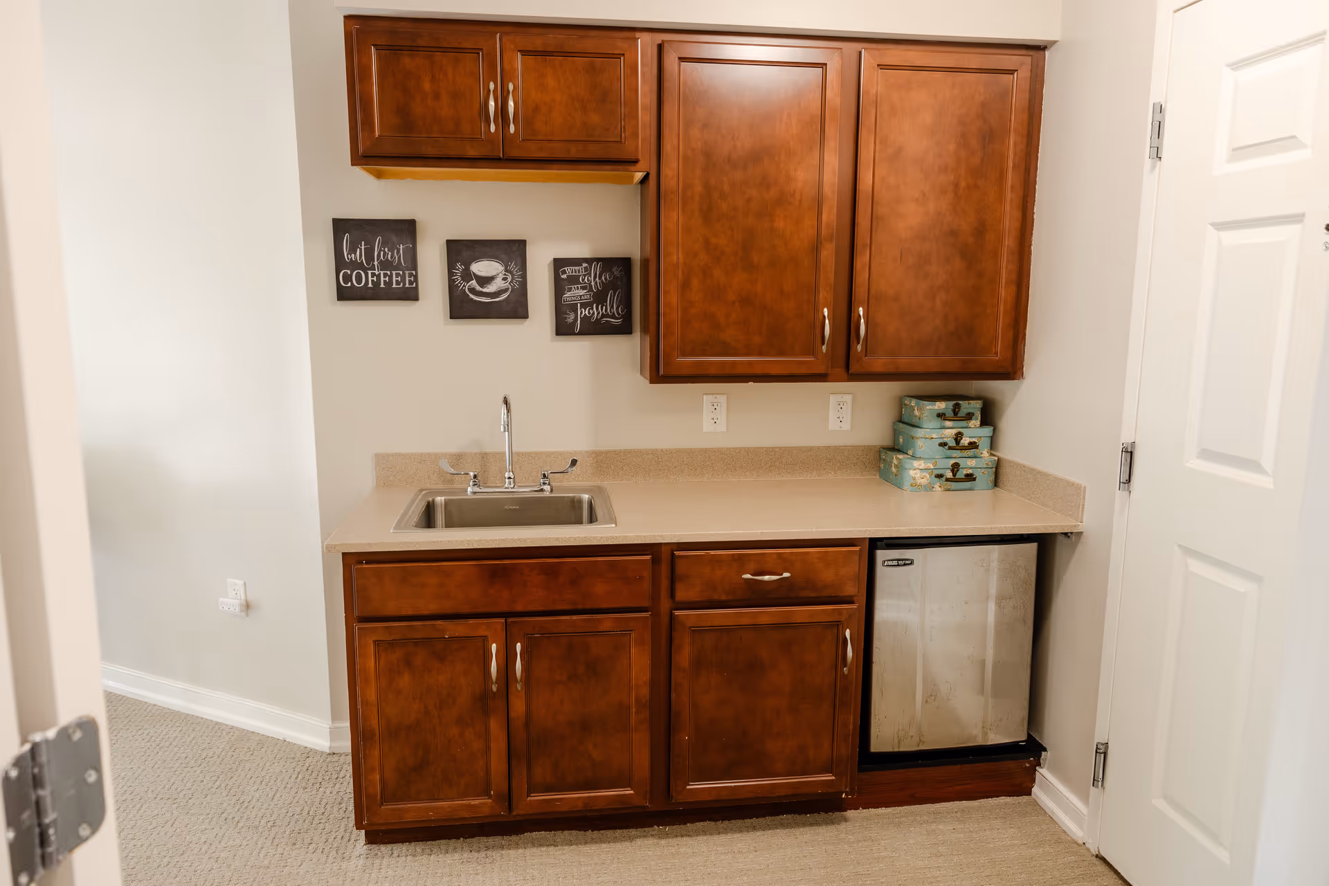 A small kitchenette area with wooden cabinets, a countertop with a sink, a mini refrigerator, and three decorative boxes stacked on the right side. Three small wall art pieces with coffee-related themes are hung above the countertop.