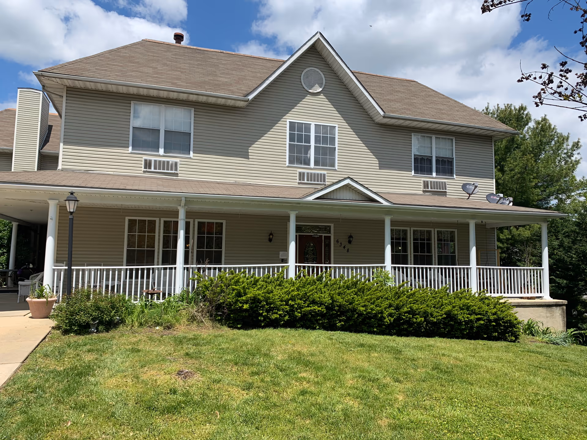 Exterior view of a two-story beige building with a large covered porch featuring white railings and columns. The building has multiple windows, a brown shingled roof, and a well-maintained green lawn in front. There are some bushes along the porch and a lamppost on the left side near a sidewalk. The sky is partly cloudy.