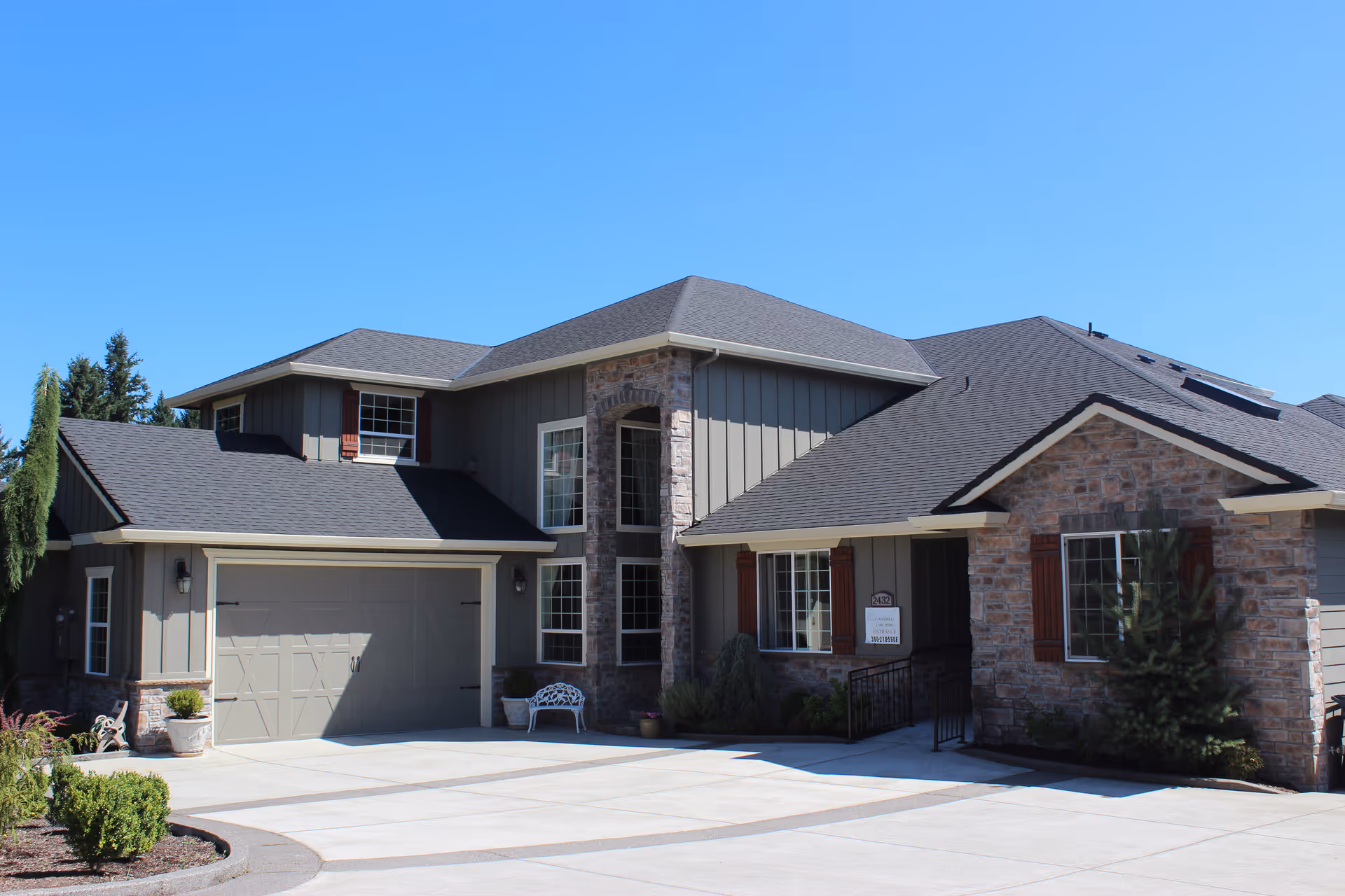 Exterior view of a modern two-story residential building with a combination of stone and wood siding, multiple windows, a garage, and a driveway under a clear blue sky.