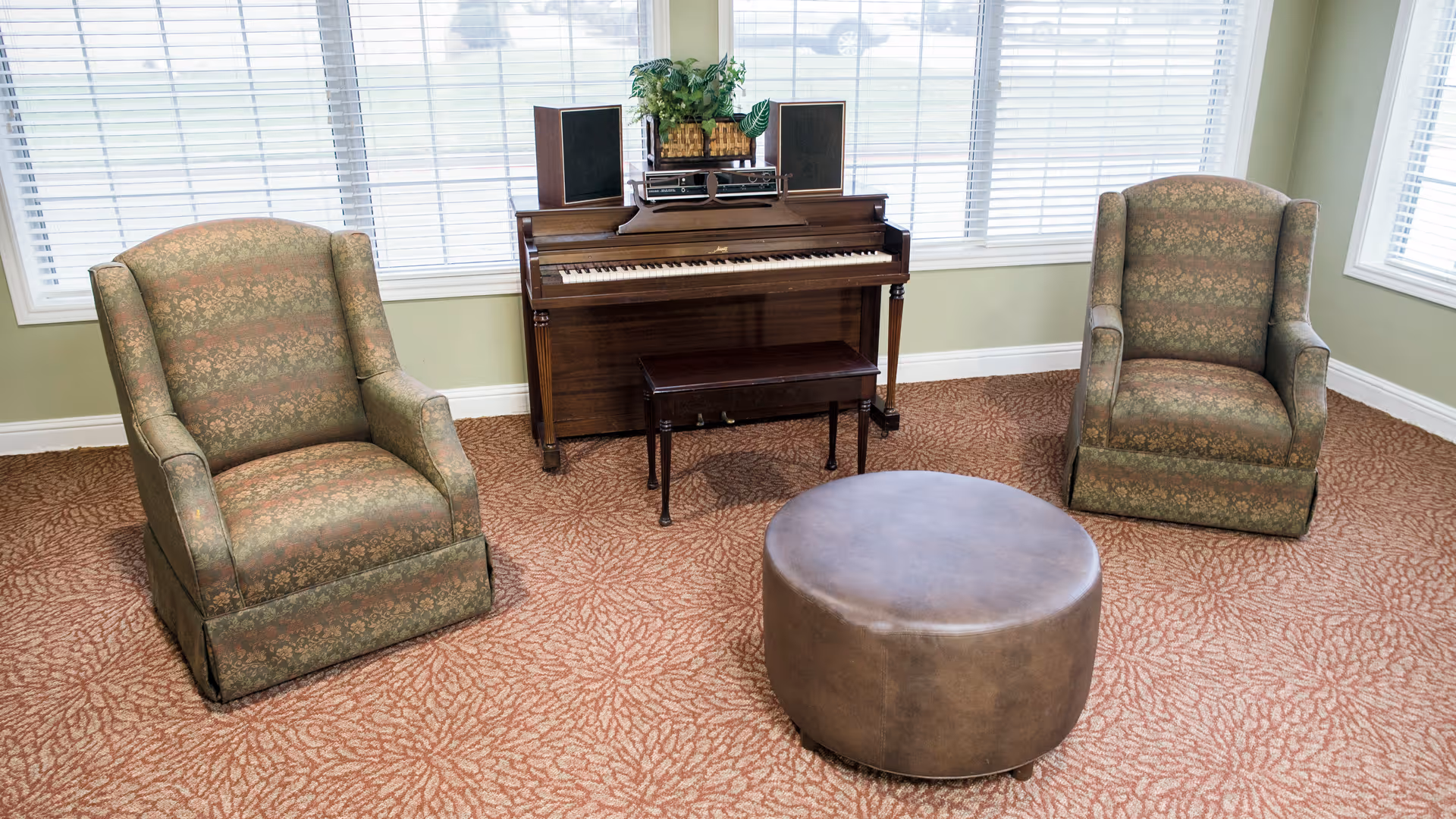 A cozy sitting area with two patterned armchairs facing a round brown ottoman, positioned in front of a wooden piano with a small bench. The piano has two speakers and a plant on top. Large windows with white blinds allow natural light into the room.