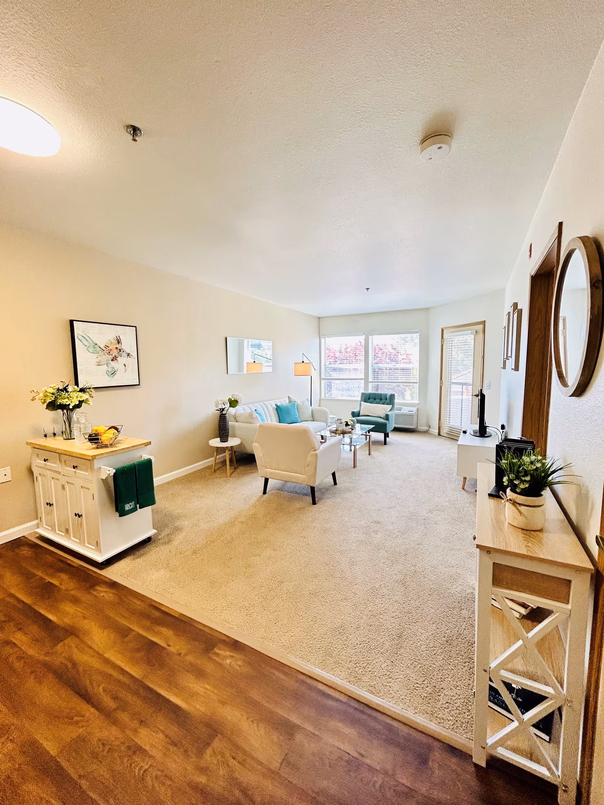A bright and spacious living room with beige carpet and light-colored walls. The room features a white armchair, a teal armchair, a white sofa with teal and white cushions, a glass coffee table, and a floor lamp. There is a small white cabinet with a wooden top holding a vase of flowers and a bowl of fruit. Large windows and a glass door let in natural light. A wooden console table with a plant and a round mirror hangs on the wall to the right.