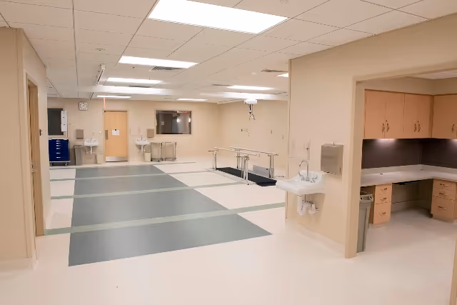 A spacious, well-lit rehabilitation room with parallel bars for walking exercises, sinks mounted on the walls, a clock, and a small office area with wooden cabinets and a countertop.