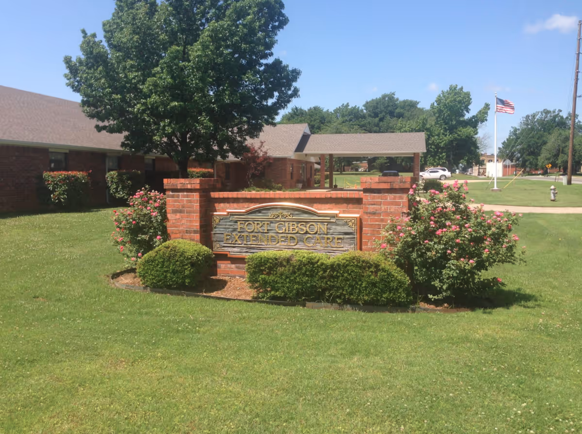 Outdoor view of Fort Gibson Extended Care facility sign made of brick and wood, surrounded by green grass, bushes, and pink flowers, with a building and trees in the background under a clear blue sky.