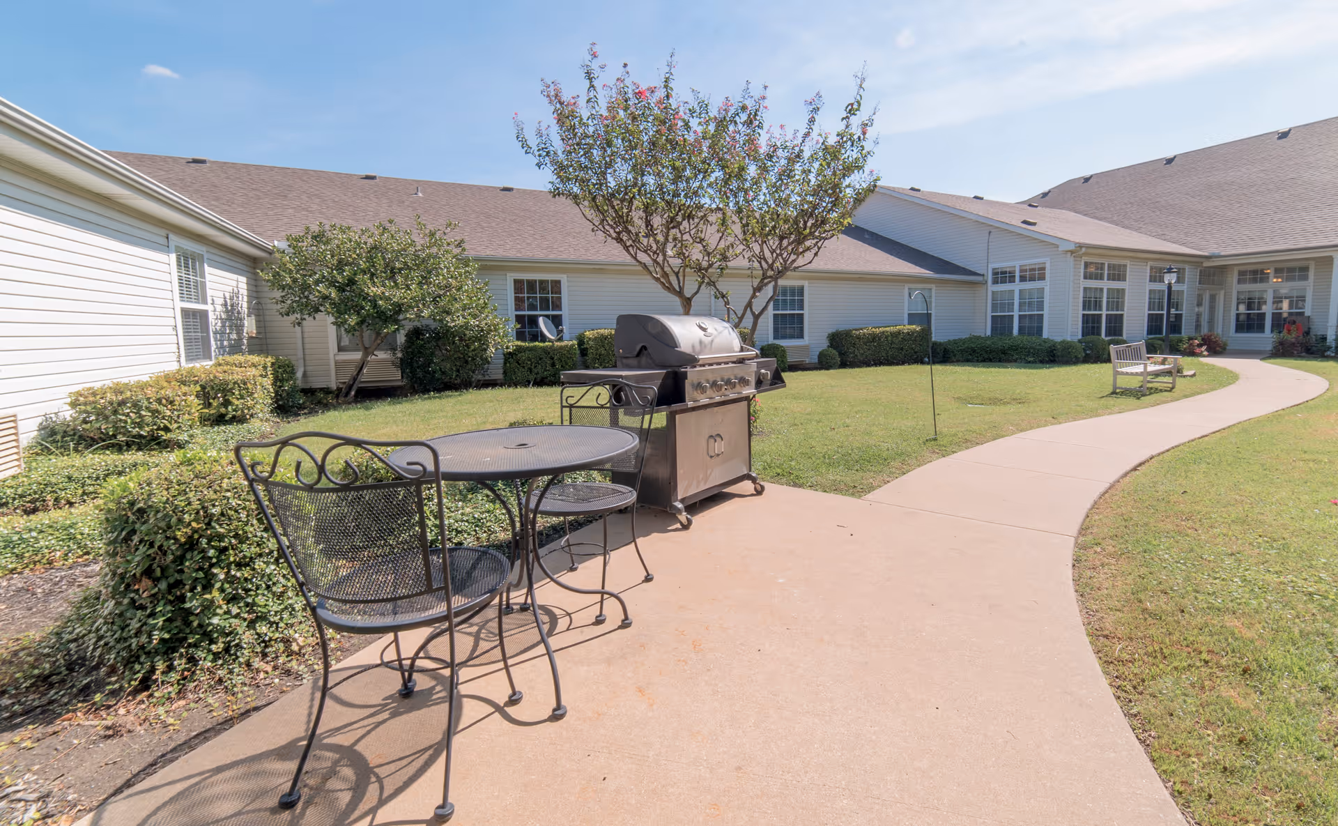 Outdoor courtyard with a paved walkway, metal patio table and chairs, a grill, benches, and single-story building surrounding a lawn.