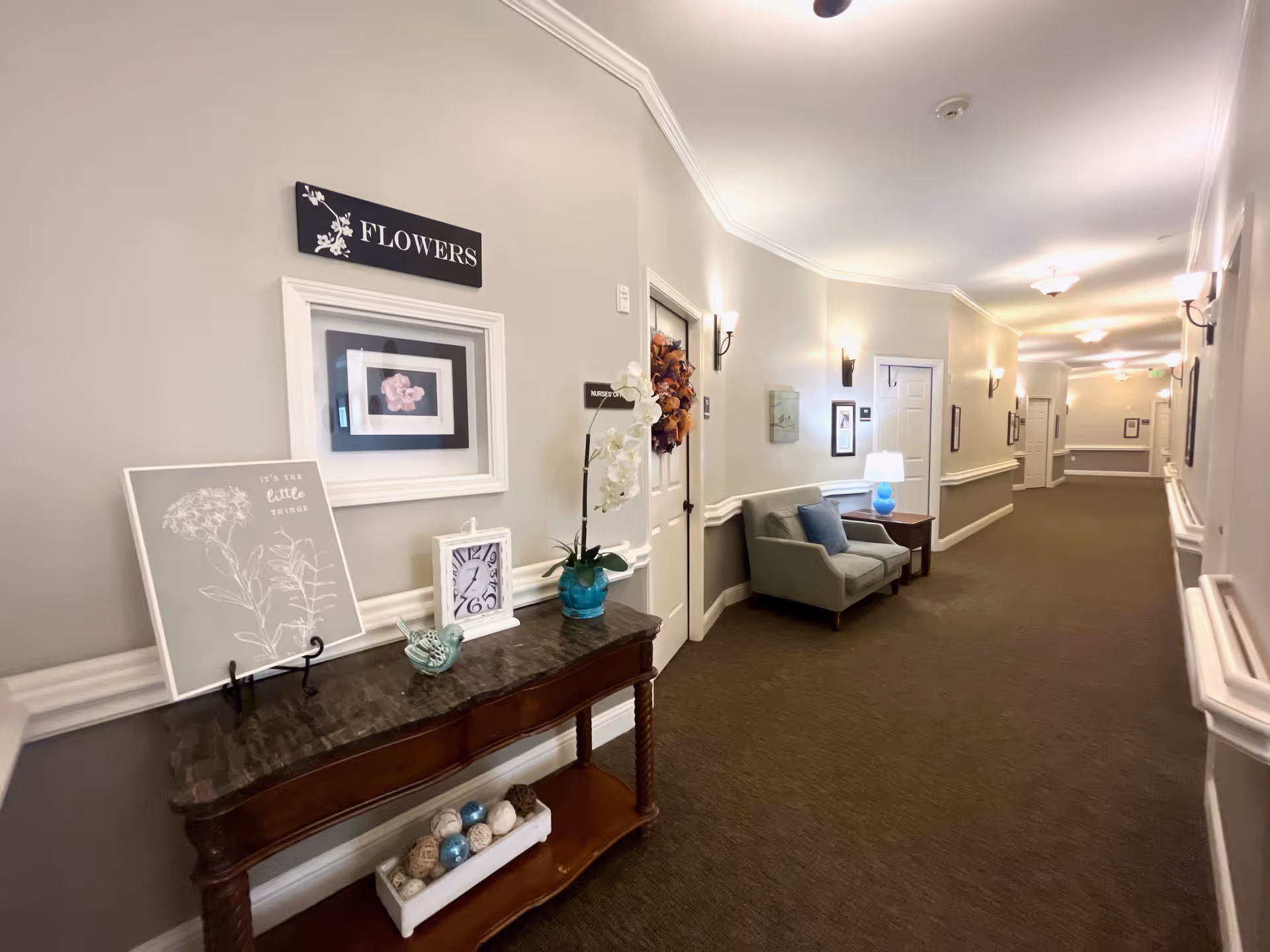 A well-lit hallway in a senior living facility with beige walls and brown carpet. On the left side, there is a wooden console table with decorative items including a clock, a small bird figurine, a framed floral print, and a potted orchid. Above the table, a sign reads 'FLOWERS'. Further down the hallway, there is a gray armchair with a blue pillow next to a small wooden side table with a blue lamp. Several doors and framed pictures line the walls, and wall sconces provide additional lighting.