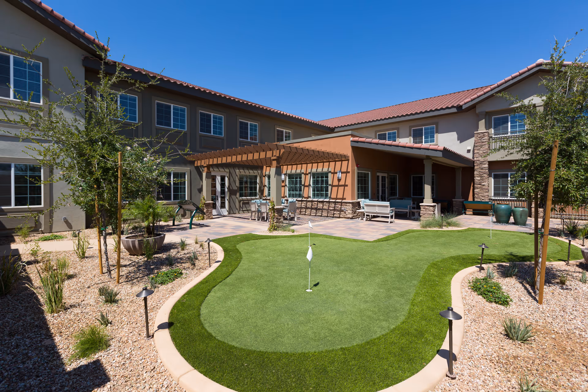 Outdoor courtyard area at The Enclave at Gilbert Senior Living featuring a small putting green surrounded by desert landscaping with rocks and plants. The building has two stories with multiple windows, a covered patio with seating, and a pergola providing partial shade.