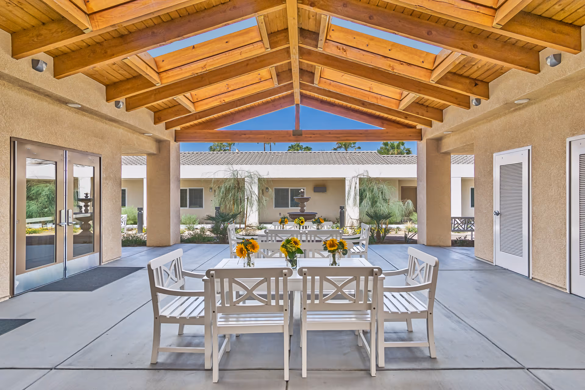 Covered outdoor seating area with white wooden tables and chairs, each table decorated with small vases of sunflowers. The area has a wooden ceiling with skylights and opens to a courtyard with plants and a water fountain in the background.