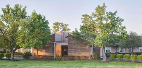 Brick healthcare building exterior with a central entrance, trees, and trimmed shrubbery.