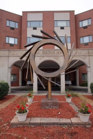Outdoor sculpture in front of a brick building with arched entrance and windows. The sculpture is circular with radiating metal beams and is surrounded by four white flower pots with red flowers placed on red mulch.