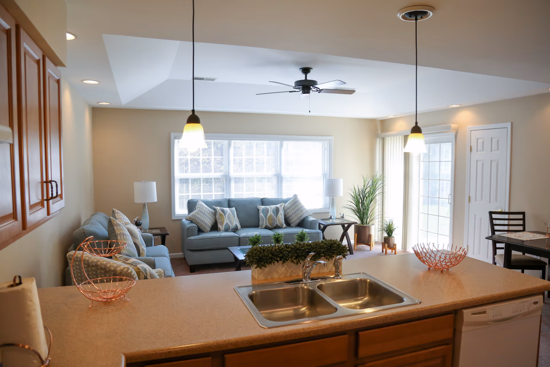 View of a bright living room and kitchen area in a senior living facility. The kitchen counter with a double sink and two pendant lights overlooks a living room with two blue sofas, patterned throw pillows, side tables with lamps, and potted plants near large windows and a glass door.