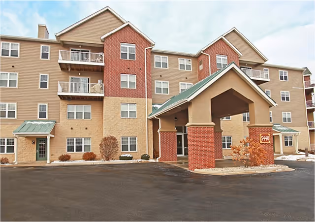 Exterior view of a multi-story senior living facility building with beige and brick facade, multiple windows, balconies, and a covered entrance with a green roof. There is a paved driveway and some small bushes around the building.
