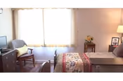 Sunlit bedroom with a bed, bedside table and lamp, dresser with a small TV, and a chair by a large curtained window.