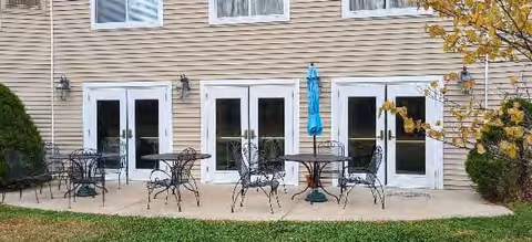 Outdoor patio area with three sets of black metal tables and chairs placed on a concrete surface in front of a beige building with multiple white-framed glass doors and windows. There is a closed blue umbrella next to one of the tables, and some greenery including bushes and a tree with yellow leaves on the right side.