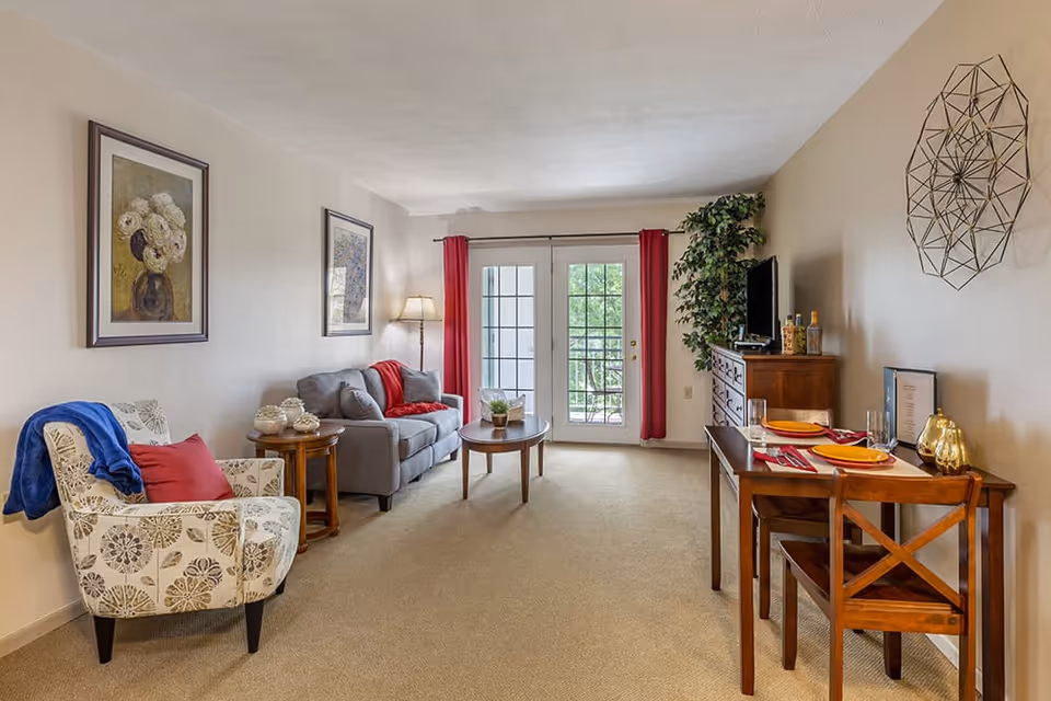 A cozy living room with a gray sofa, a floral patterned armchair with a blue throw, a wooden coffee table, and a small dining table set for two. The room has beige walls adorned with framed artwork and a decorative metal wall piece. French doors with red curtains lead to an outdoor balcony with greenery visible outside.