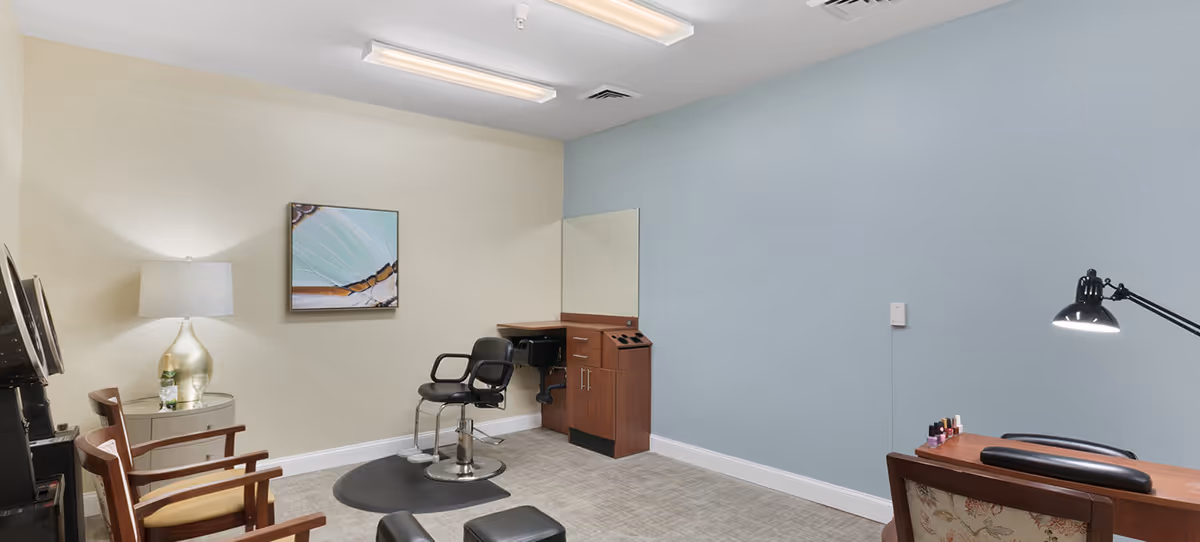 A small salon room with a hair washing station, a salon chair, two wooden chairs, a side table with a lamp and a bottle, a wall mirror, a nail care station with a lamp and nail polish bottles, and light blue and beige walls.