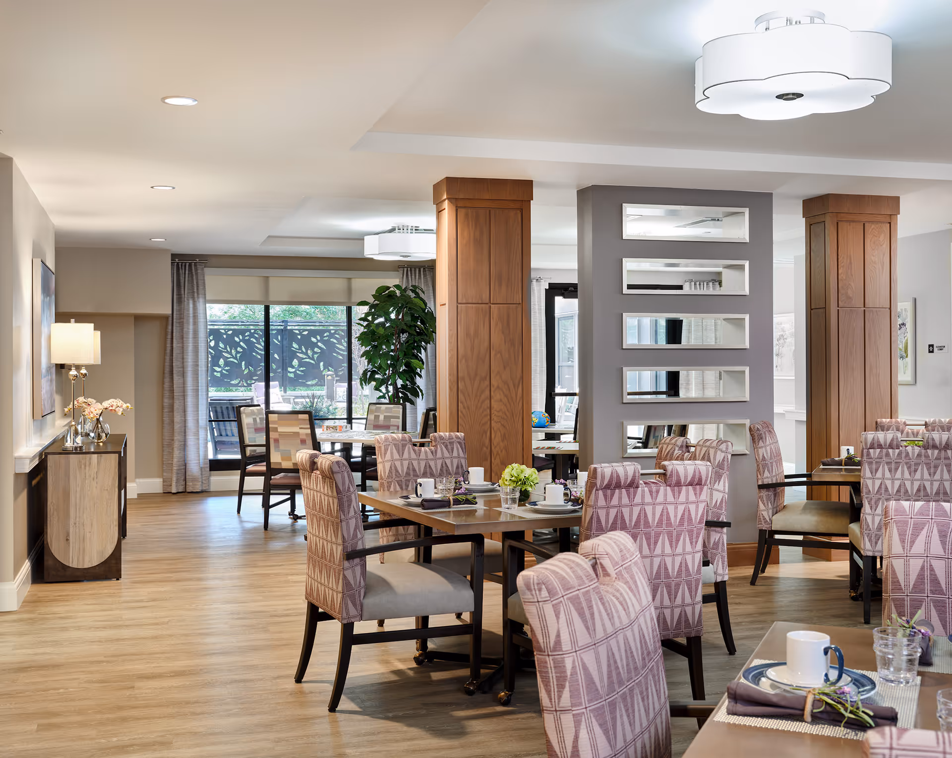 Bright assisted-living dining room with patterned upholstered chairs around wooden tables, decorative wood pillars, and large windows.