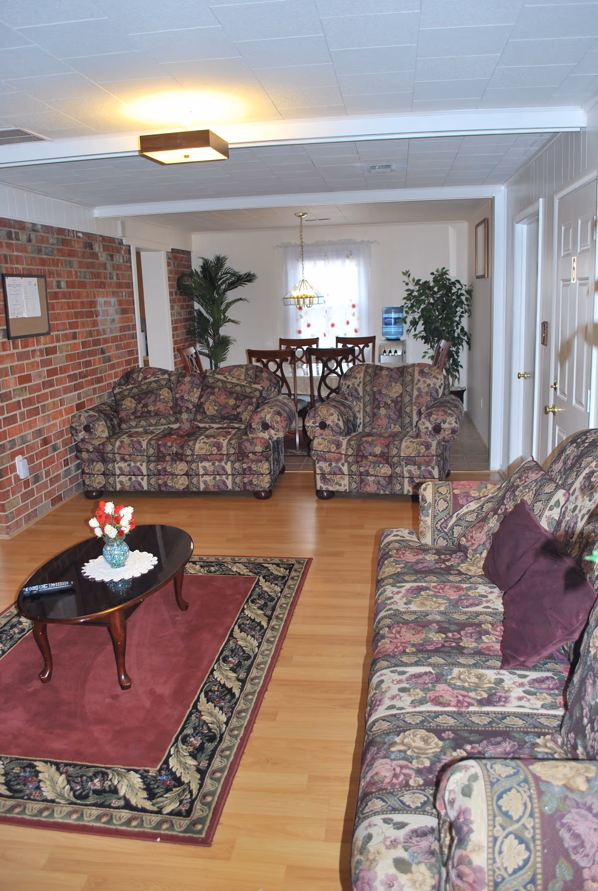 A living room with floral patterned sofas and armchairs arranged around a dark wooden coffee table on a red and floral area rug. The room has a brick accent wall on the left and a wooden floor. In the background, there is a dining area with a wooden dining table and chairs, a hanging light fixture, and some potted plants near a window with sheer curtains.