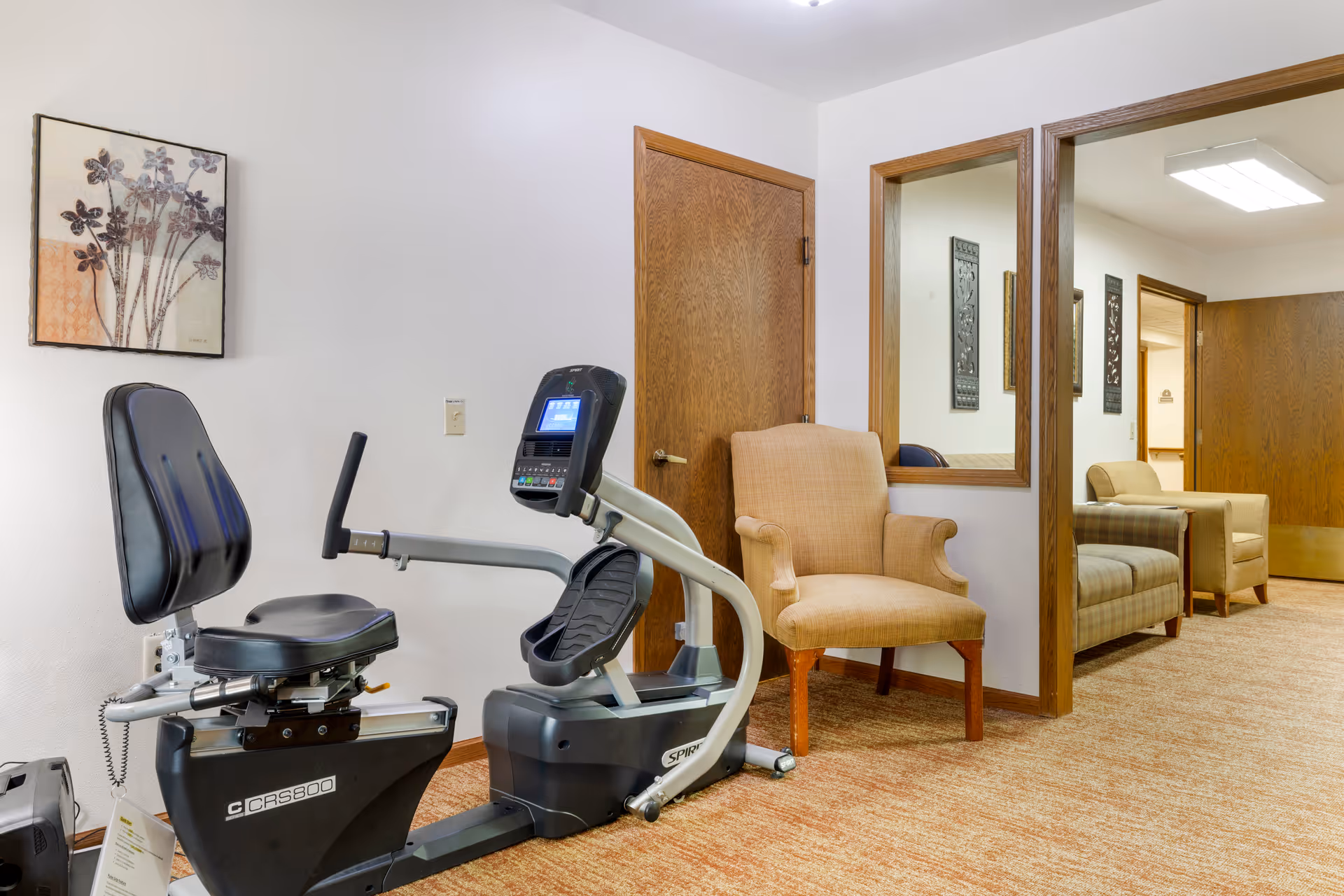 An interior common area with a recumbent exercise bike, upholstered chairs, and a doorway leading to a seating area.