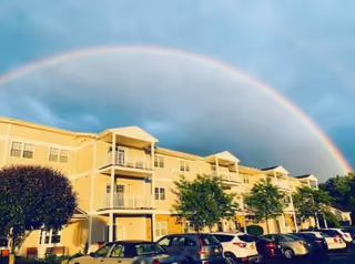 A yellow multi-story residential building with balconies and several parked cars in front under a cloudy sky with a vibrant rainbow arching over the building.