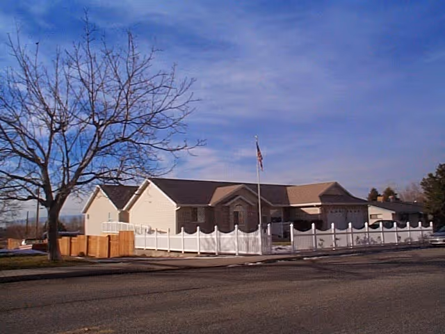Single-story residential building with beige siding and a dark roof, surrounded by a white picket fence. There is a flagpole with an American flag in front of the building. A leafless tree is visible on the left side, and the sky is clear with some clouds.