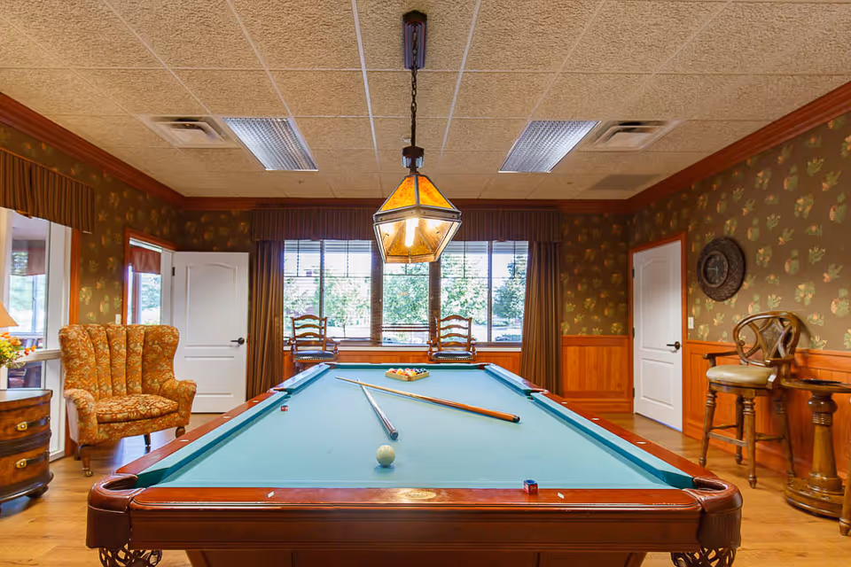 Interior view of a recreational room with a pool table in the center. The pool table has a green felt surface with pool balls arranged in a triangle and two cues resting on it. The room features patterned wallpaper, wooden wainscoting, a hanging light fixture above the table, an upholstered armchair on the left, two wooden chairs near a window with blinds, and a wooden stool with a backrest on the right. There are two white doors on either side of the window.