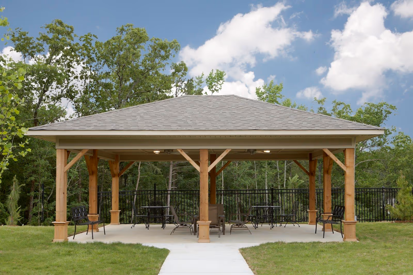A wooden gazebo with a shingled roof situated on a concrete patio surrounded by grass. Inside the gazebo are several metal chairs and tables. Trees and a black metal fence are visible in the background under a partly cloudy sky.