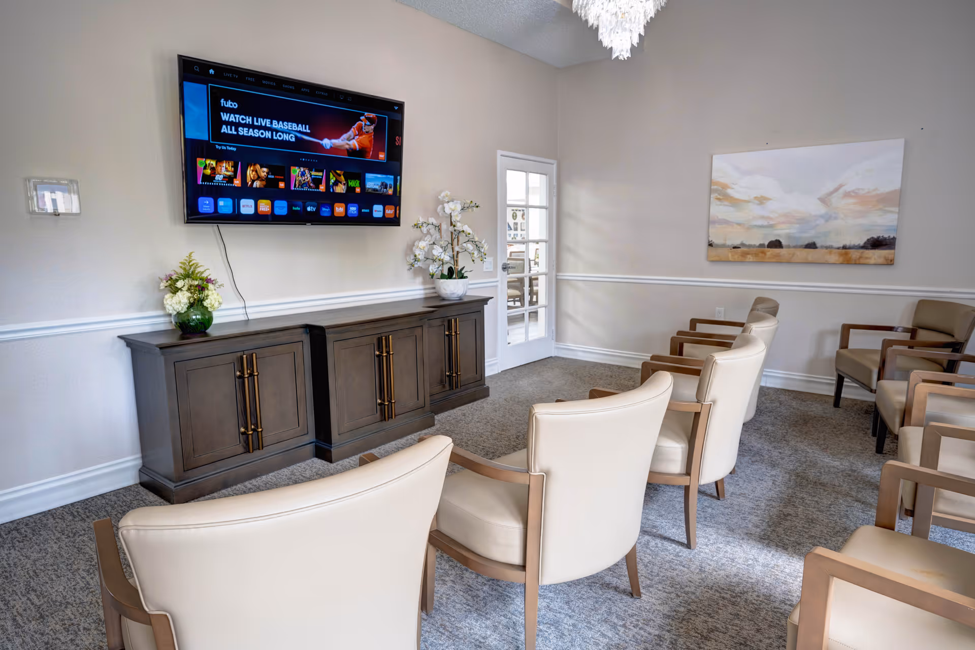 A cozy common area with beige armchairs arranged in rows facing a wall-mounted flat-screen TV displaying a sports streaming service. Below the TV is a dark wooden cabinet with decorative plants on top. The room has light-colored walls, a carpeted floor, a chandelier, and a landscape painting on the wall.
