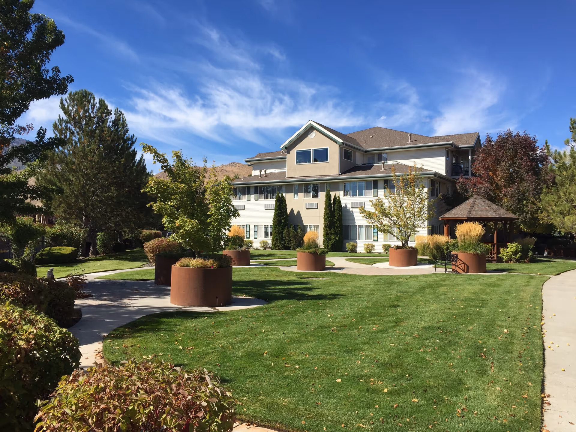 A three-story retirement community building with beige and white exterior walls, surrounded by a well-maintained lawn and landscaped garden. There are several large round planters with small trees and shrubs, a paved walkway, and a wooden gazebo on the right side. The sky is clear with some wispy clouds.