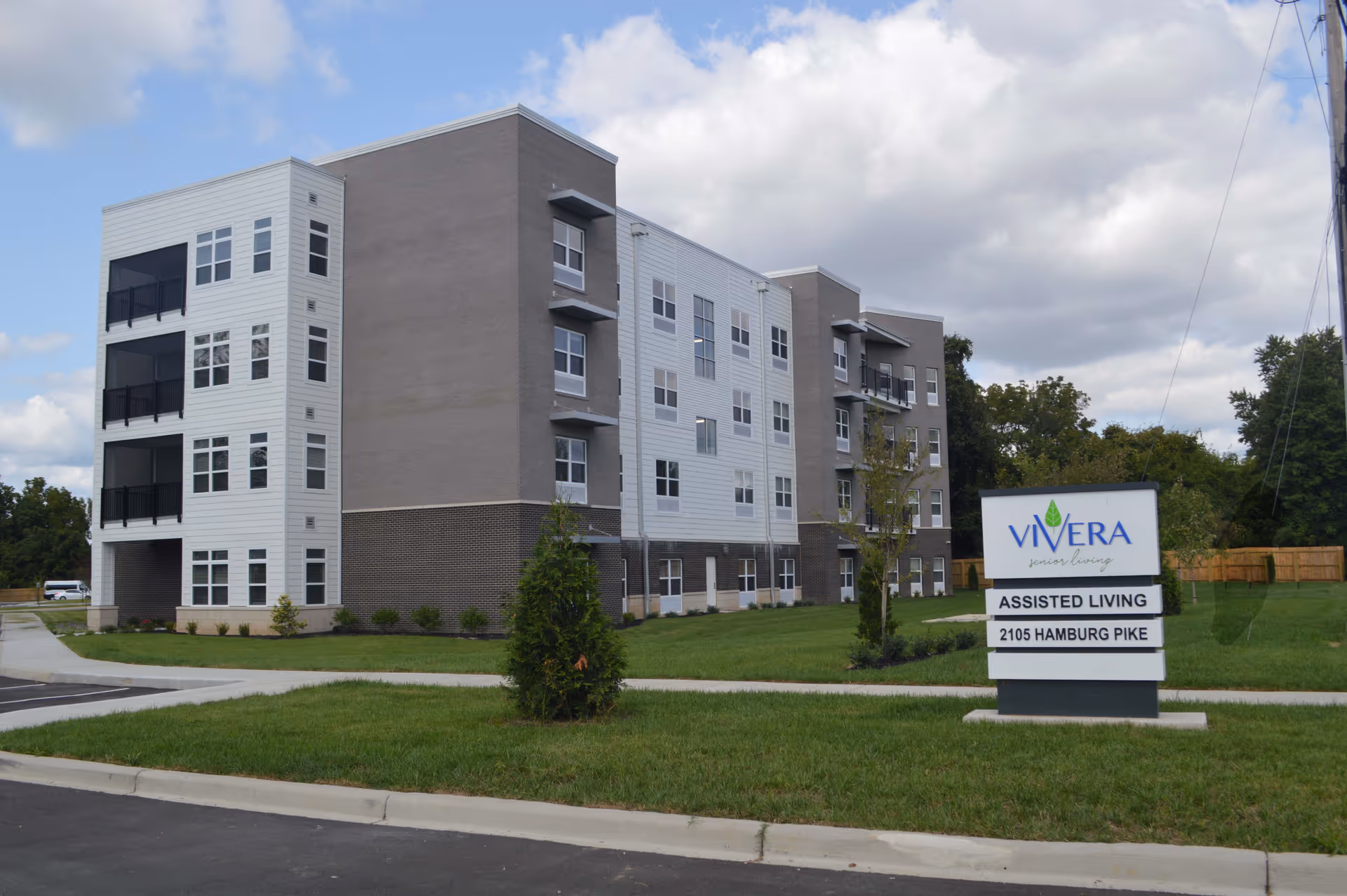 Exterior view of a modern multi-story assisted living building with a sign in front that reads Vivera Senior Living, Assisted Living, 2105 Hamburg Pike. The building has a combination of white and gray exterior walls with multiple windows and balconies. The surrounding area includes a well-maintained lawn and a sidewalk.