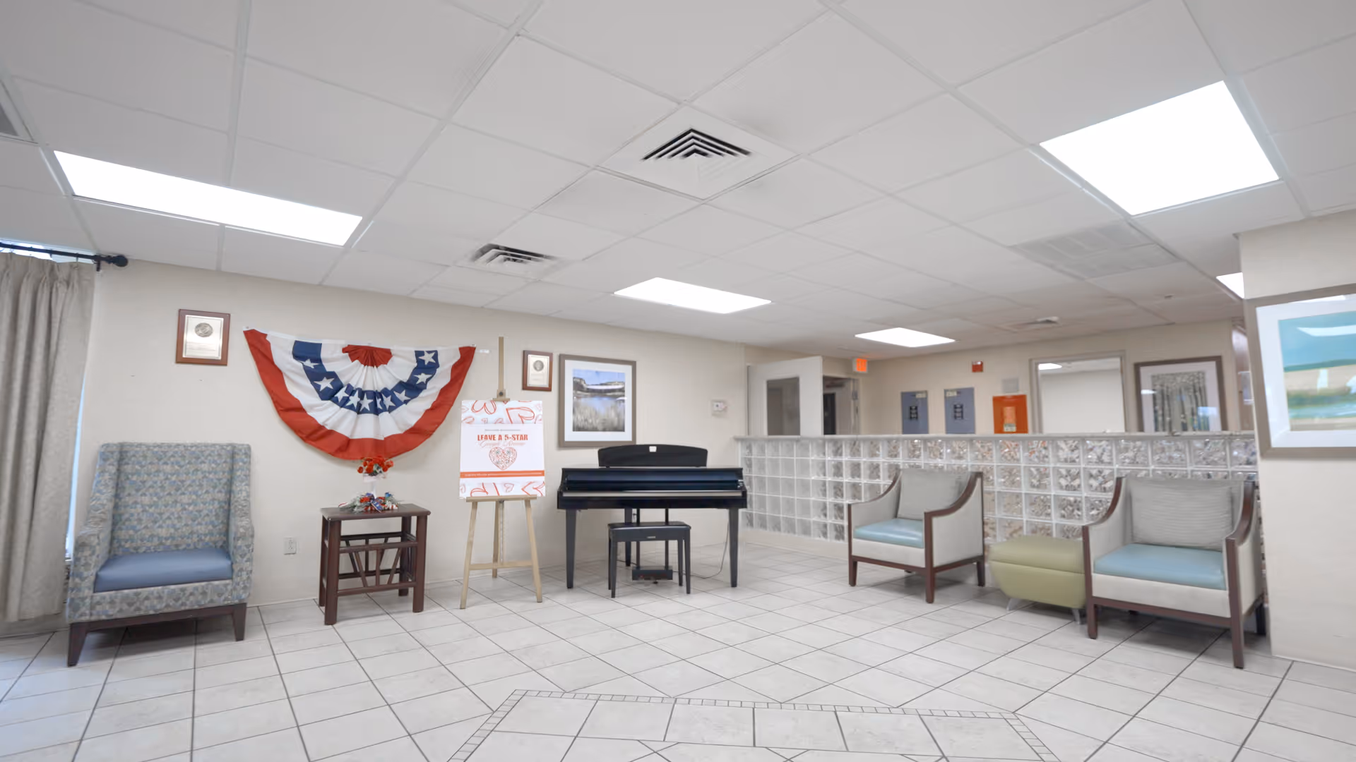 A bright communal lounge with seating, a piano, patriotic bunting on the wall, and a glass-block divider.