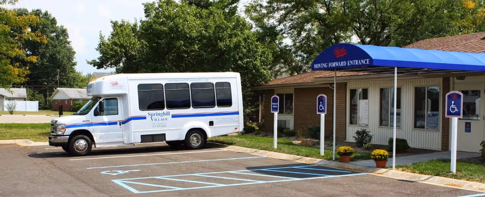 A Springhill Village shuttle van parked outside a building entrance with a blue canopy and marked accessible parking spaces.