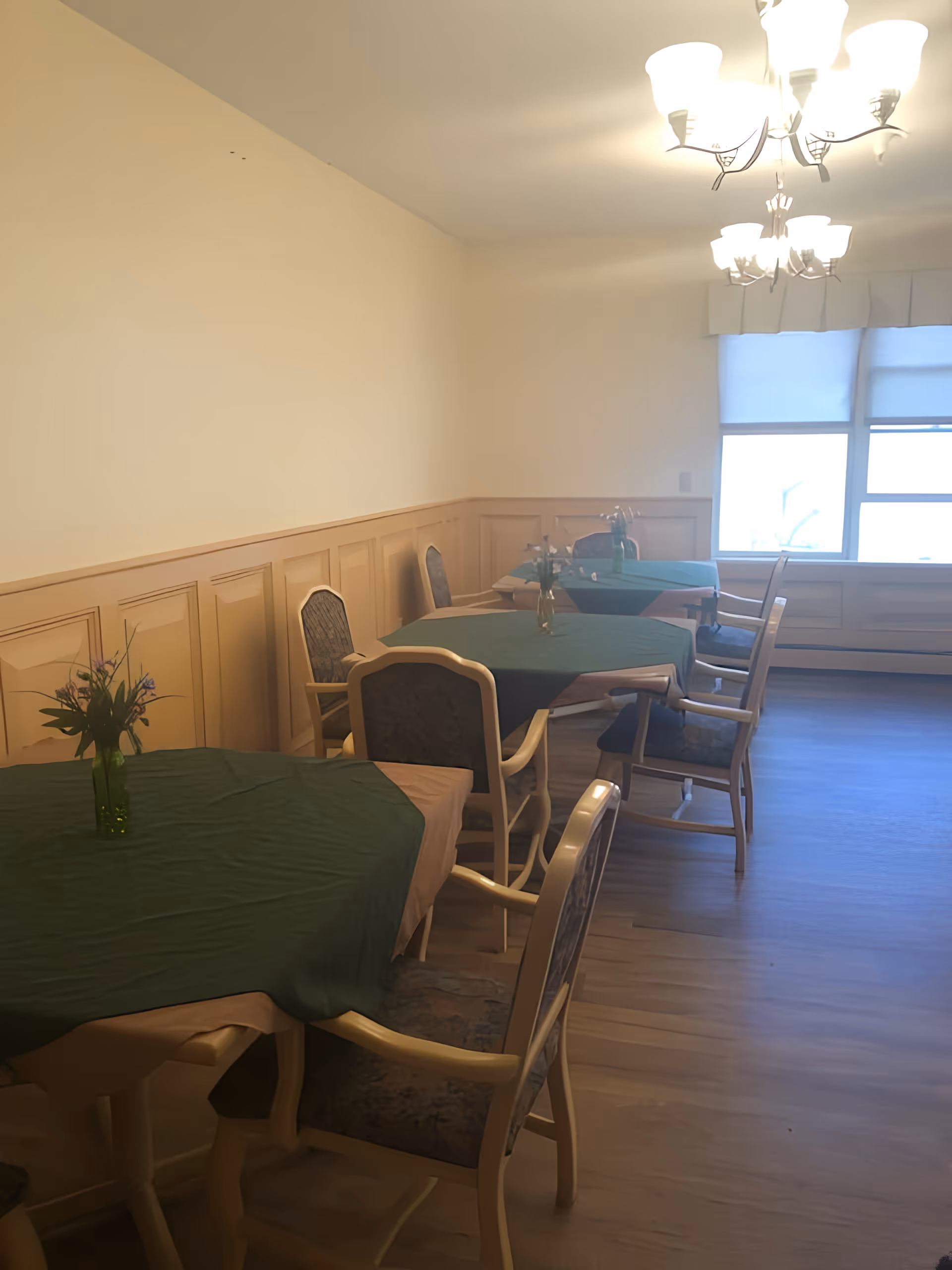 Dining room with multiple round tables draped in green tablecloths, chairs, floral centerpieces, wood paneling and a lit chandelier by a window.