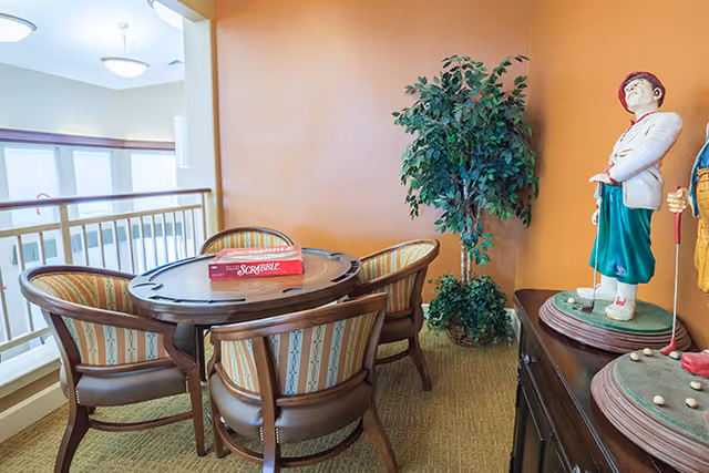 A cozy corner in a senior living facility with a round wooden table surrounded by four upholstered chairs. A Scrabble board game is placed on the table. There is a green potted plant against an orange wall, and two decorative golf player figurines on a dark wooden cabinet nearby. The area overlooks a bright space with large windows and a railing.