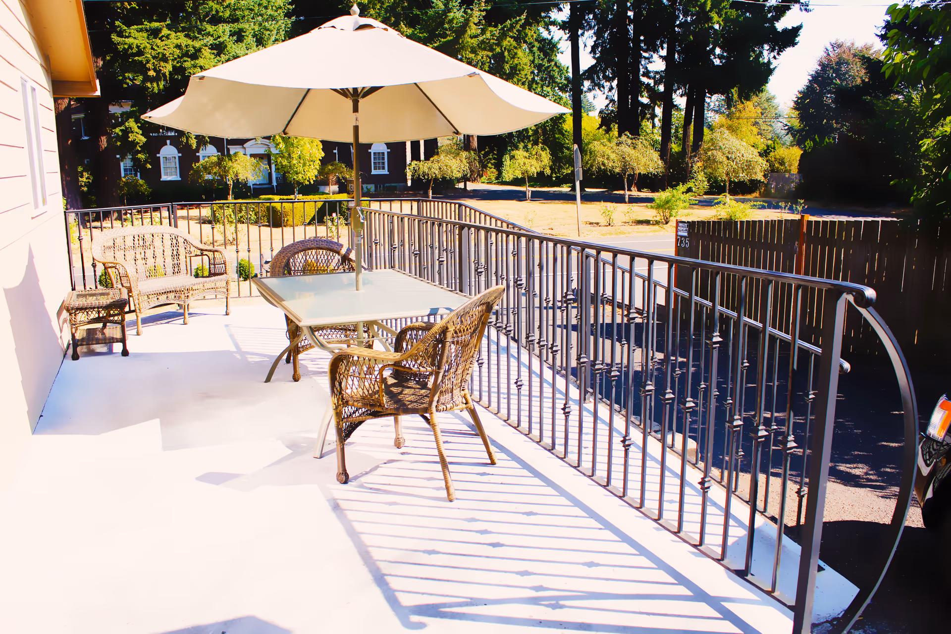 Outdoor patio area with a glass-top table, four wicker chairs, a wicker loveseat, and a large white umbrella providing shade. The patio is enclosed by a black metal railing and overlooks a parking area and trees in the background.