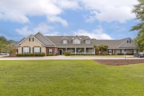 Front exterior view of Sumter Terrace Assisted Living facility, showing a large single-story building with a combination of brick and siding, multiple windows, a covered porch with white railings, and a well-maintained lawn in the foreground under a partly cloudy sky.