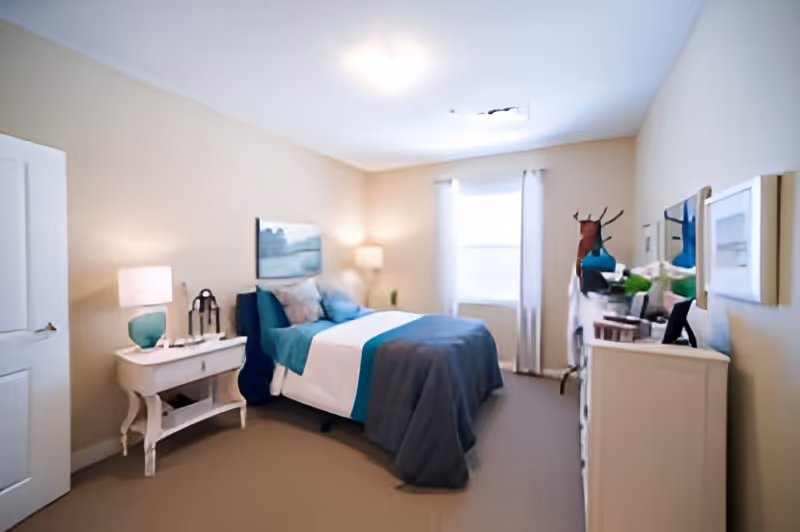 A cozy bedroom in a senior living facility with a neatly made bed featuring teal and gray bedding. The room has beige walls, a window with white curtains, a white nightstand with a lamp and decorative items, and a white dresser with a mirror and various decorations.
