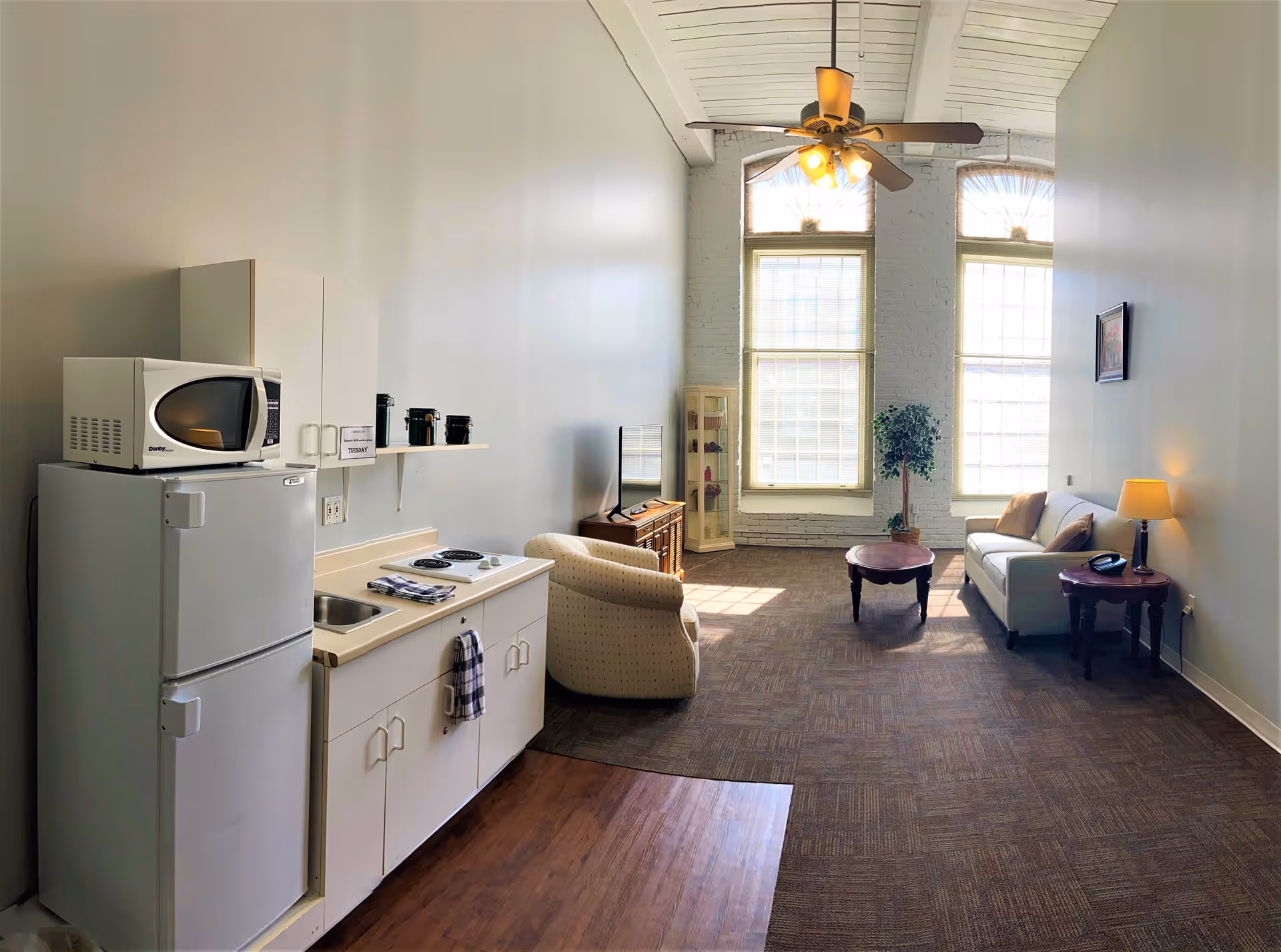 A bright living area with a small kitchenette on the left, including a refrigerator, microwave, sink, and stovetop. The room has high ceilings with a ceiling fan and two large windows letting in natural light. There is a beige armchair, a wooden TV stand with a flat-screen TV, a white sofa with pillows, a round wooden coffee table, a side table with a lamp and phone, a tall plant, and a glass display cabinet near the windows.