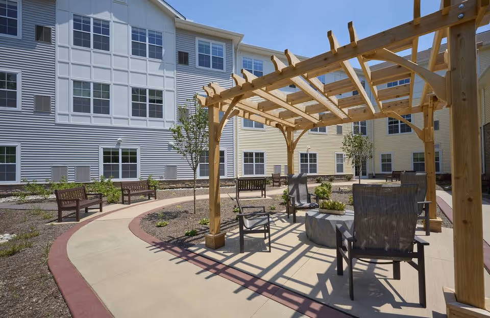 Outdoor courtyard area at The Heritage Of Green Hills featuring a wooden pergola with chairs and a small table underneath, surrounded by a paved walking path, benches, young trees, and a multi-story building with many windows in the background under a clear blue sky.
