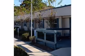 Exterior view of a single-story building with a covered walkway, small trees, and shrubs along the sidewalk under a clear sky.