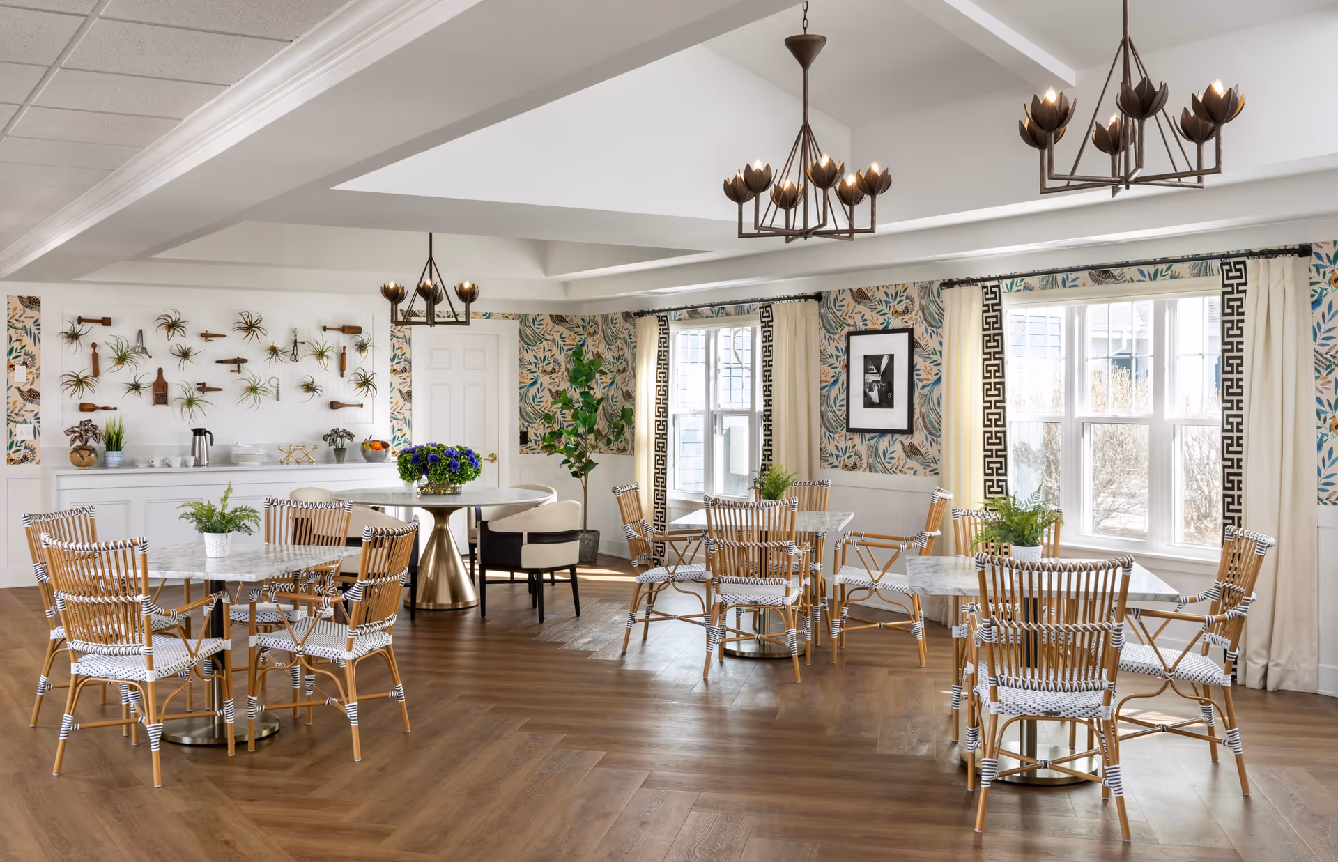 A bright and airy dining room with multiple marble-top tables surrounded by wicker chairs with white and black woven seats. The room features large windows with cream curtains bordered by a black geometric pattern, floral wallpaper, and wooden flooring. Two modern chandeliers with flower-shaped light fixtures hang from the ceiling. A sideboard with plants, a coffee pot, and cups is visible against one wall, which is decorated with mounted air plants and wooden utensils.