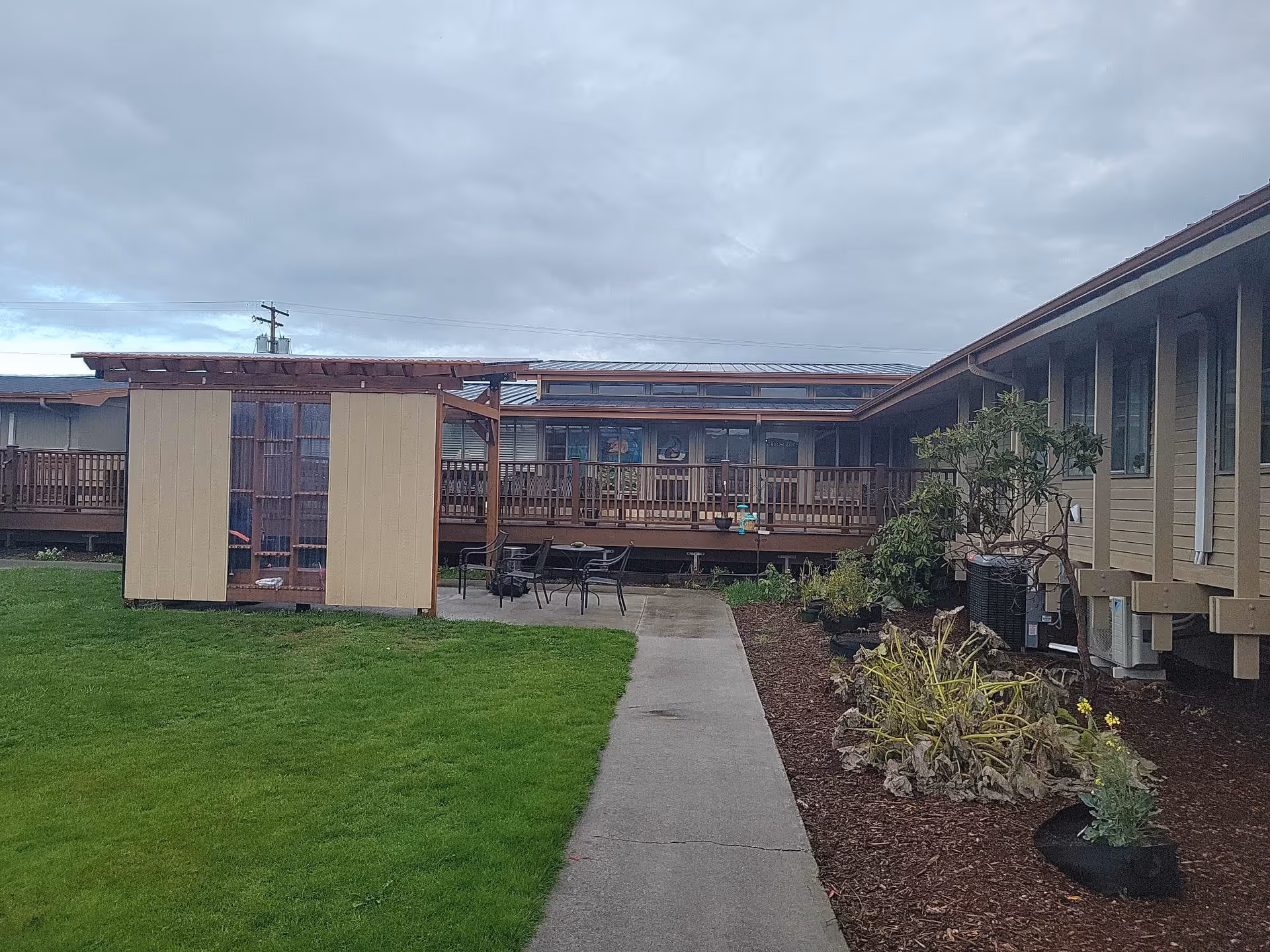 Outdoor view of a senior living facility courtyard with a concrete walkway leading to a small patio area with chairs and a table. There is a wooden structure on the left side and a garden bed with plants on the right side. The building surrounds the courtyard and has windows and a wooden deck with railings.