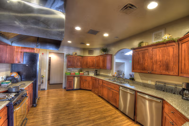 A spacious kitchen with wooden cabinets, granite countertops, stainless steel appliances including a refrigerator, dishwasher, and stove. The kitchen has wooden flooring and recessed lighting in the ceiling. There is an open archway leading to another room with visible chairs and tables.