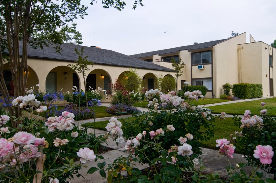 View of a senior living facility building with beige walls and a dark roof, featuring arched walkways and multiple windows. The foreground shows a garden with blooming pink and white flowers, green shrubs, and a paved walkway. The sky is clear with a single bird flying.