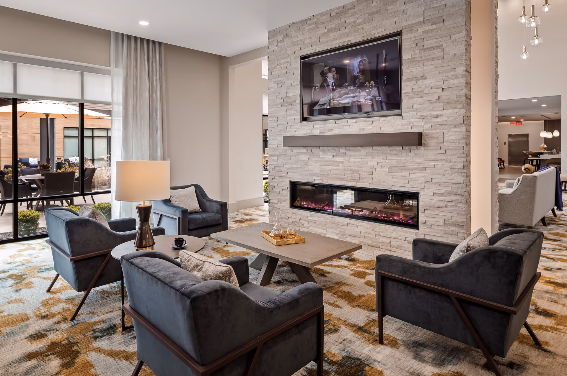 A modern living room area with four dark blue armchairs arranged around a rectangular wooden coffee table. A table lamp is on a small round side table between two chairs. The room features a stone accent wall with a built-in electric fireplace and a mounted flat-screen TV above it. Large windows reveal an outdoor patio with seating and umbrellas. The carpet has a patterned design in shades of beige and brown.